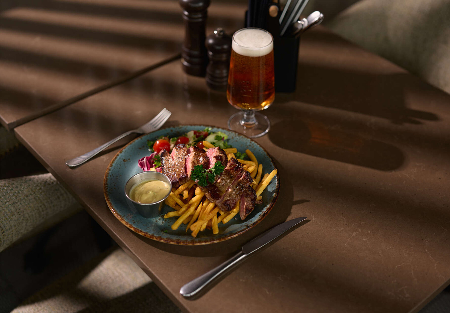 Tallrik med skivad biff, pommes frites, sallad och en skål med sås, serverad med ett glas öl.